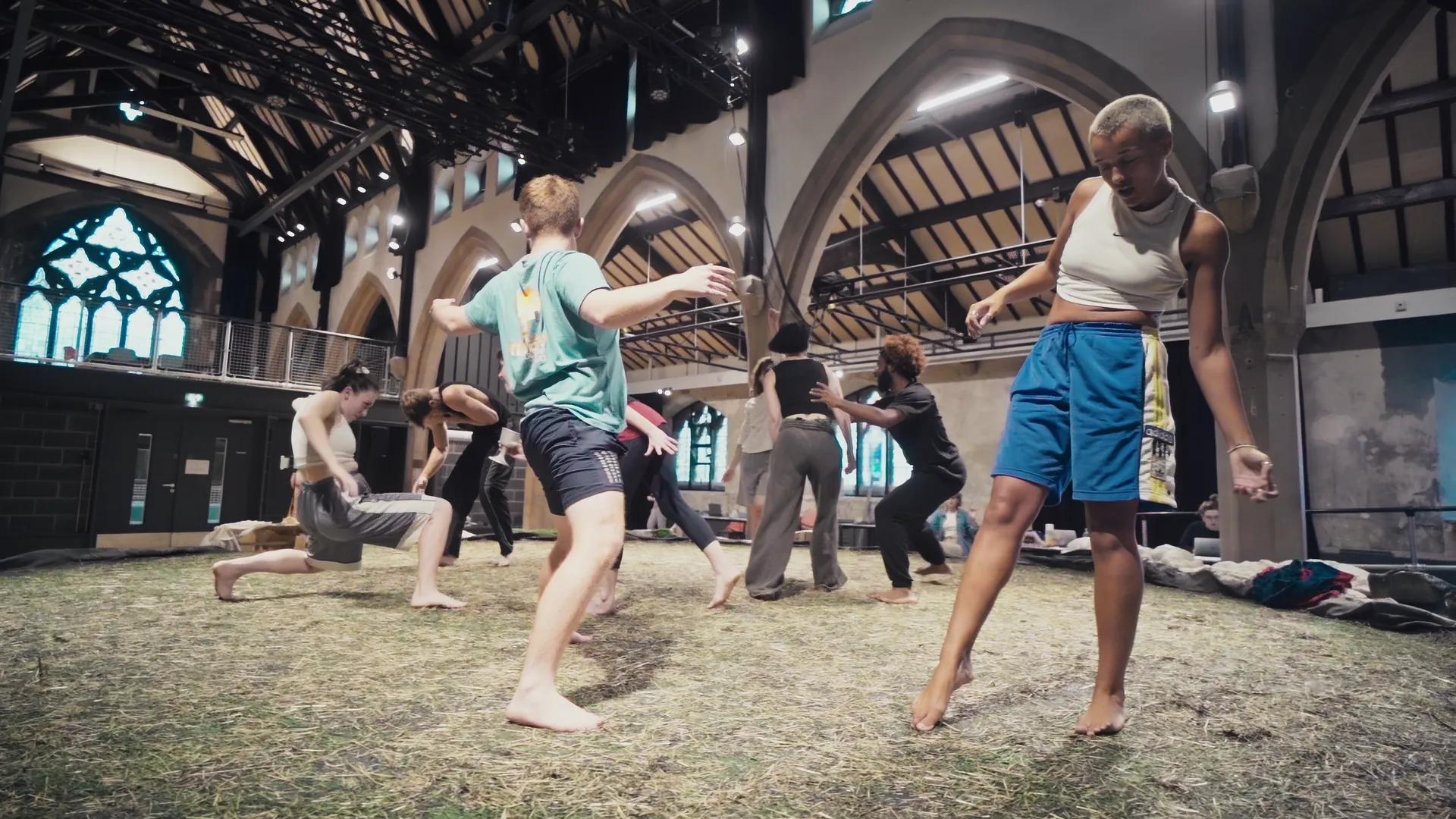 Fran Wen OLION ensemble company barefoot mid warm-up on the hay-strewn rehearsal floor under arched stone windows.