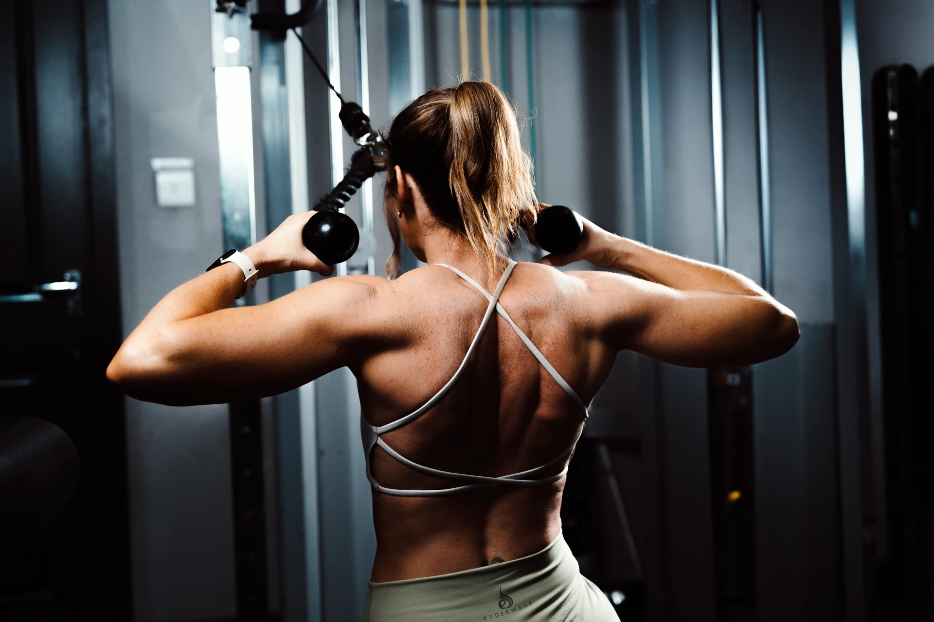 Cable rope pull from behind — back anatomy detail, gym light wash. Editorial fitness photography by Mona Digital.