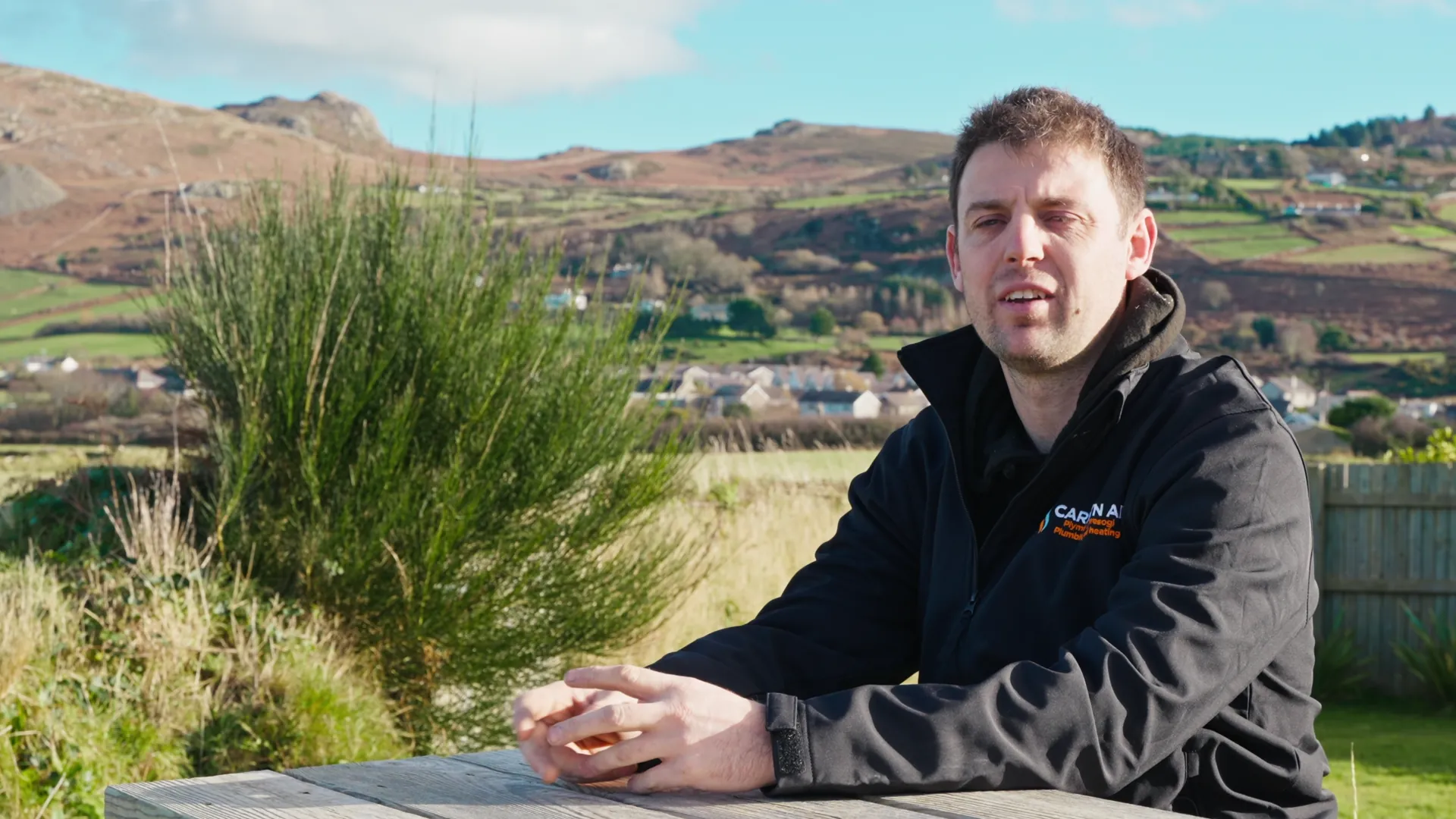 Carwyn interviewed seated at a wooden picnic table with Yr Eifl mountains and Llŷn farmhouses behind — Menter Môn case-study film.