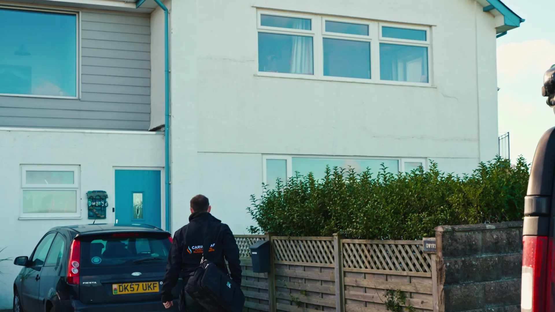 Carwyn arriving at a customer's home in branded jacket carrying his tool bag — documentary moment from the Menter Môn case-study film.