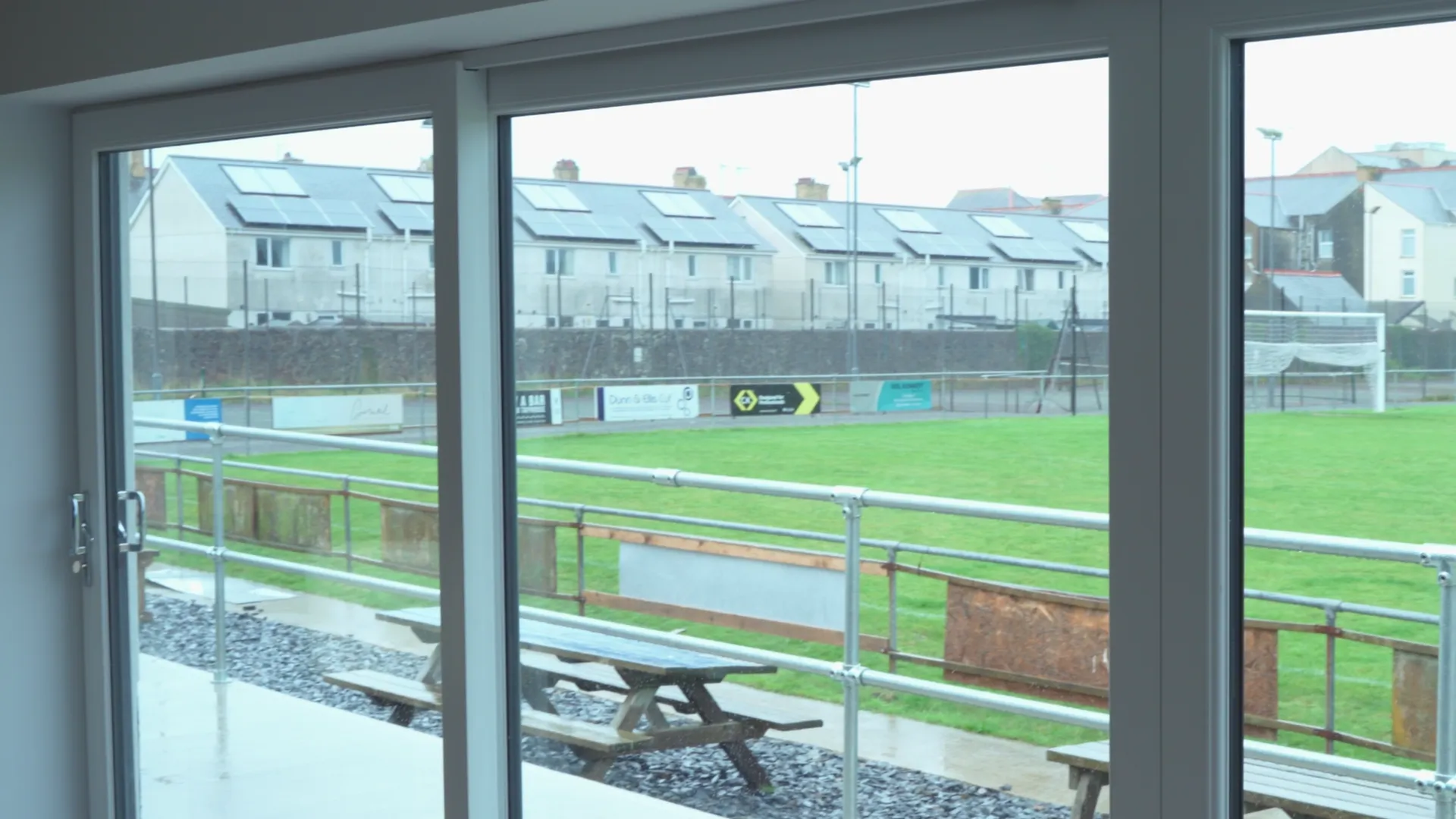 Through-glass view of the CPD Pwllheli pitch from the clubhouse — Menter Môn film about Llŷn Peninsula community football.