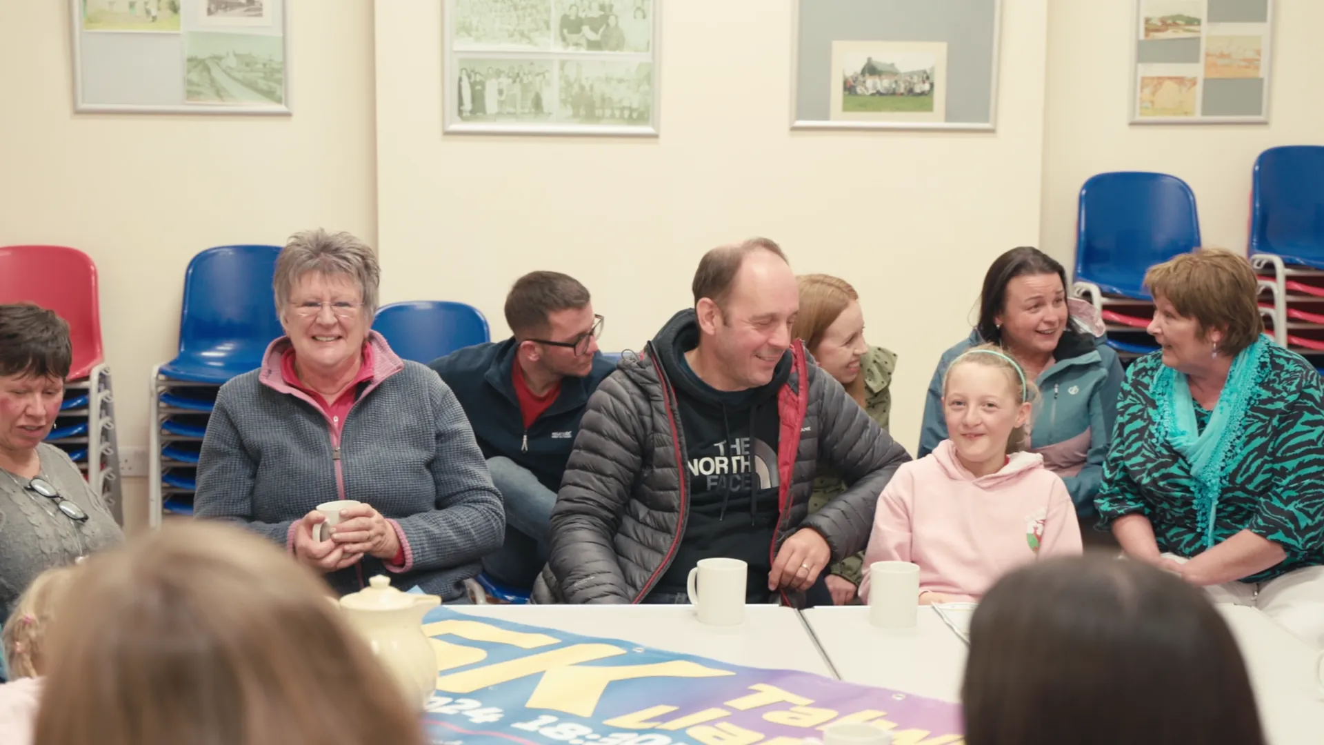 Ffrindiau Llanbedrgoch community group meeting around a table at the village hall, smiling — Menter Môn film about volunteer-run village life on Anglesey.