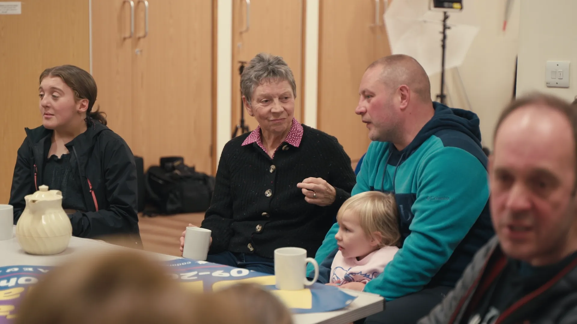 Elder community member in conversation with a young father holding a toddler — intergenerational community meeting in Llanbedrgoch village hall.