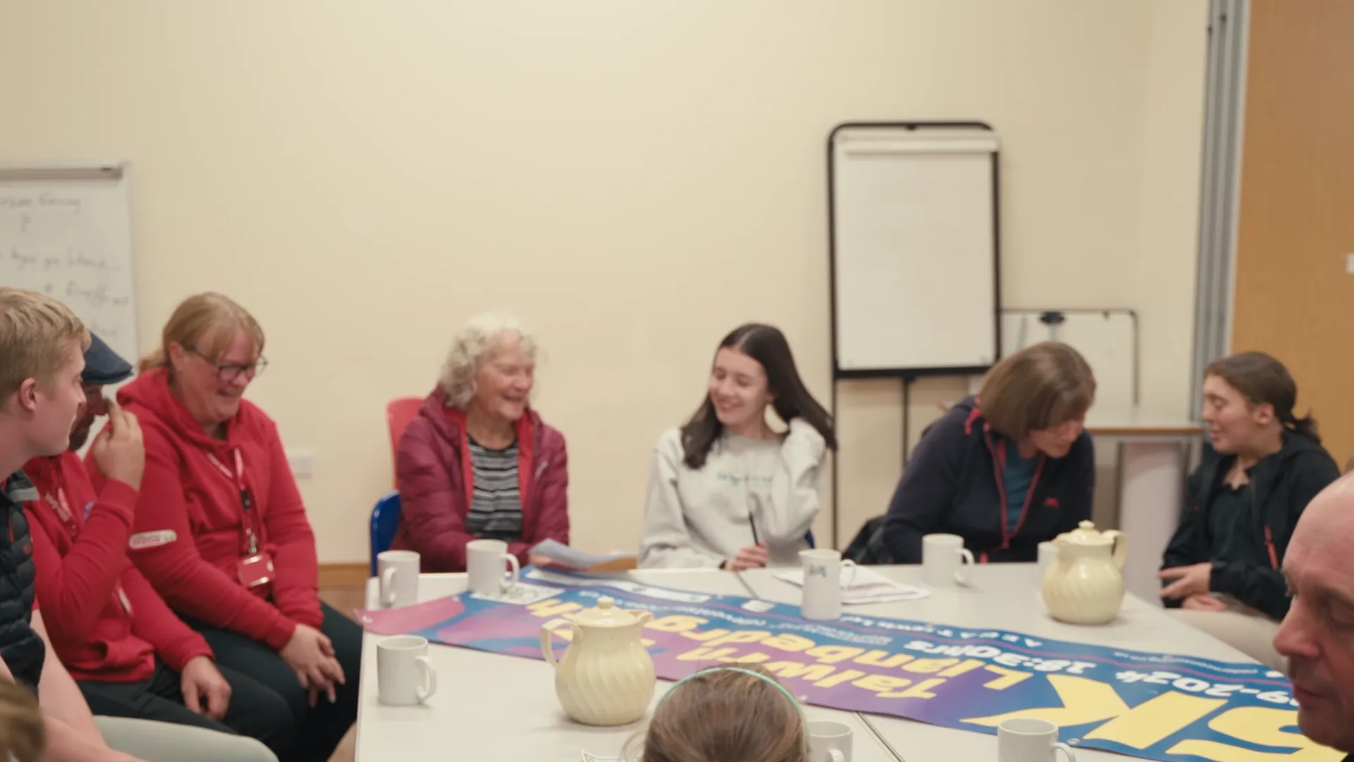 Mixed-ages community meeting at Llanbedrgoch village hall — teenagers, parents, grandparents around the table planning events for Ffrindiau Llanbedrgoch.
