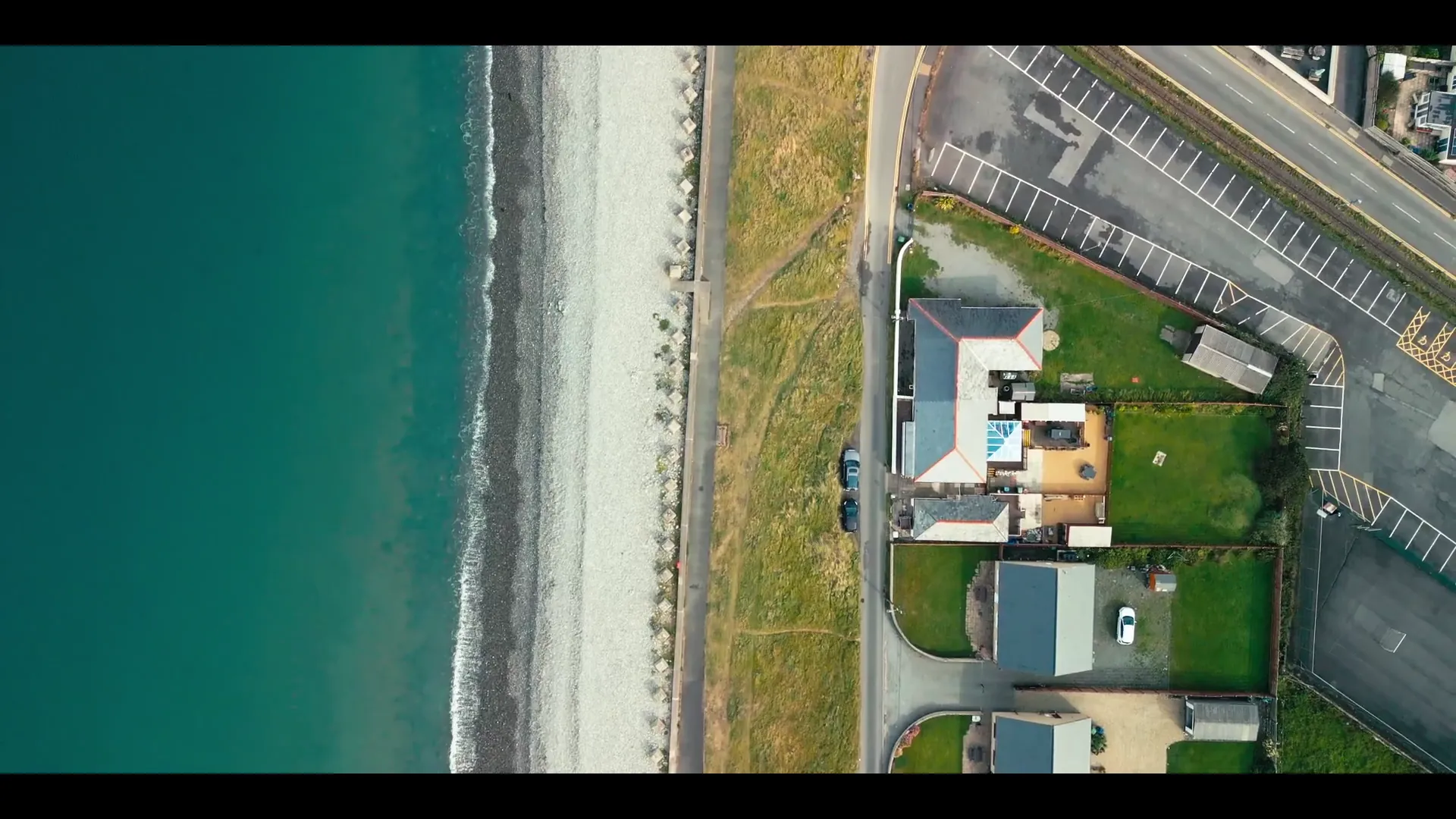 Top-down drone aerial of Welsh coastal terraces with the railway line running parallel to a beach of turquoise water — Mona Digital showreel.